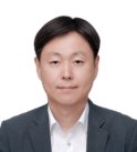A korean man with short brown hair wearing a white collared shirt and a dark grey suit jacket gives a slight smile to the camera in front of a white background.